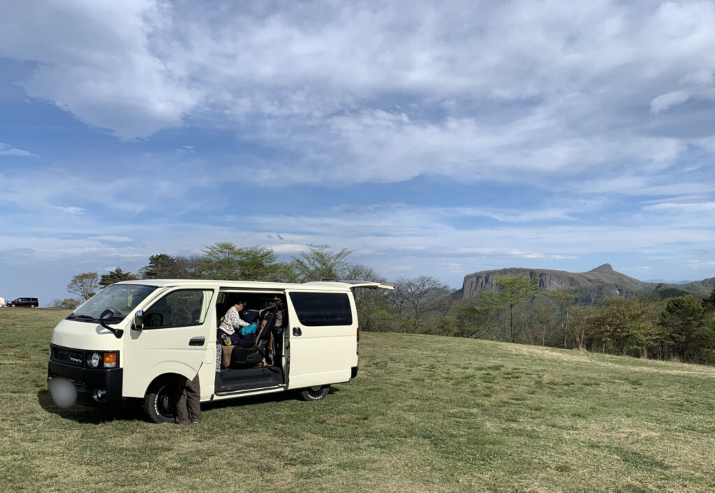 ライトイエローのハイエース200系を山が見える草原に停め、スライドドアを開けてキャンプを楽しんでいる様子