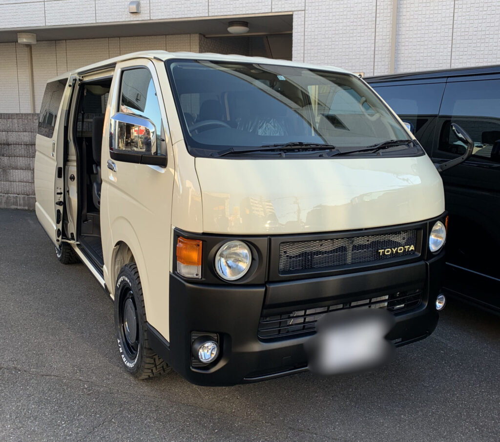 ライトイエローのハイエース200系カスタム。丸目ヘッドライト、ブラックバンパー、オフロードタイヤを装着したフロント。